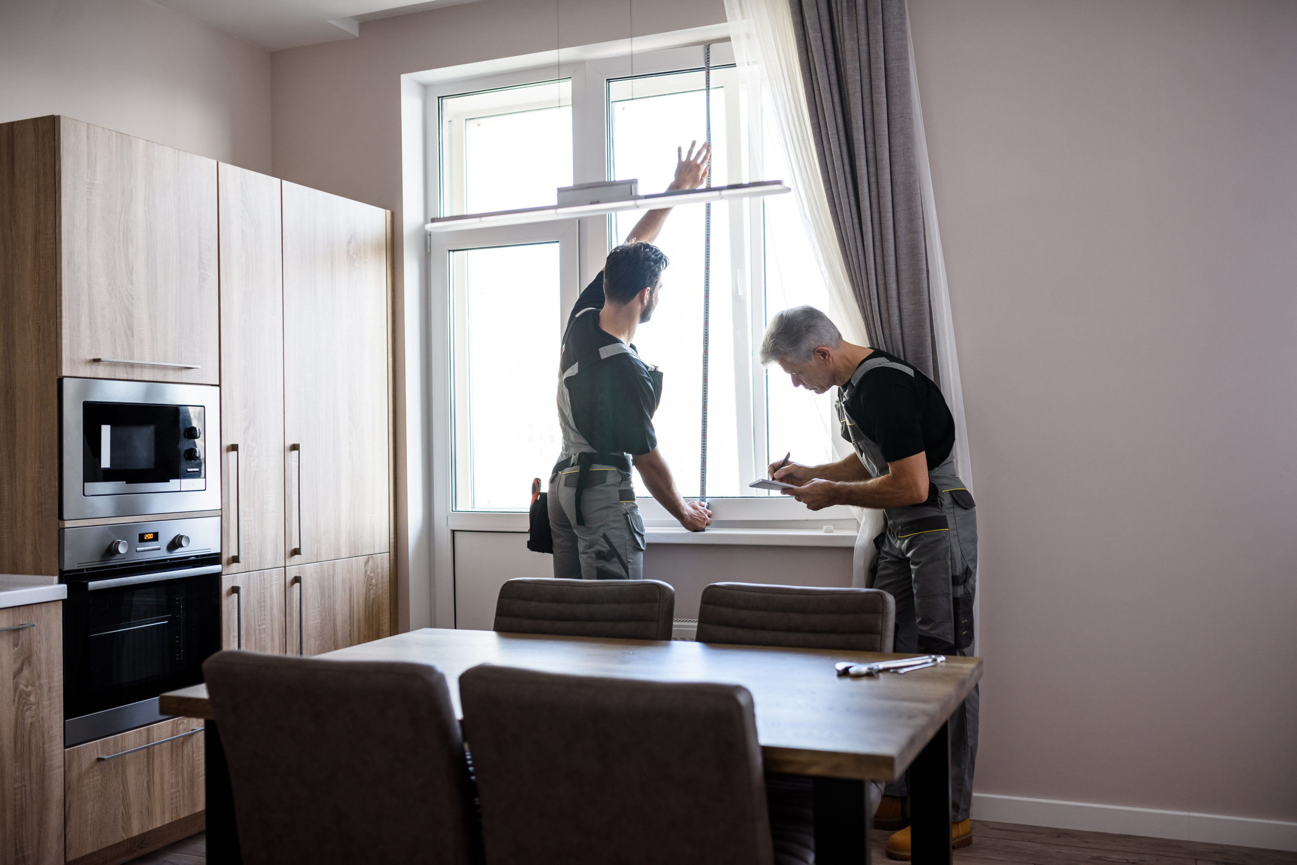 Young professional worker in uniform using tape measure, measuring window for installing blinds, while his aged colleague making notes. Construction and maintenance concept